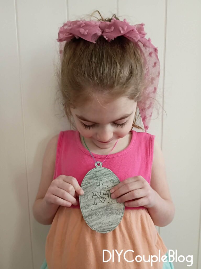 girl showing the back of a catholic printable miraculous medal craft necklace colored in "silver" crayon