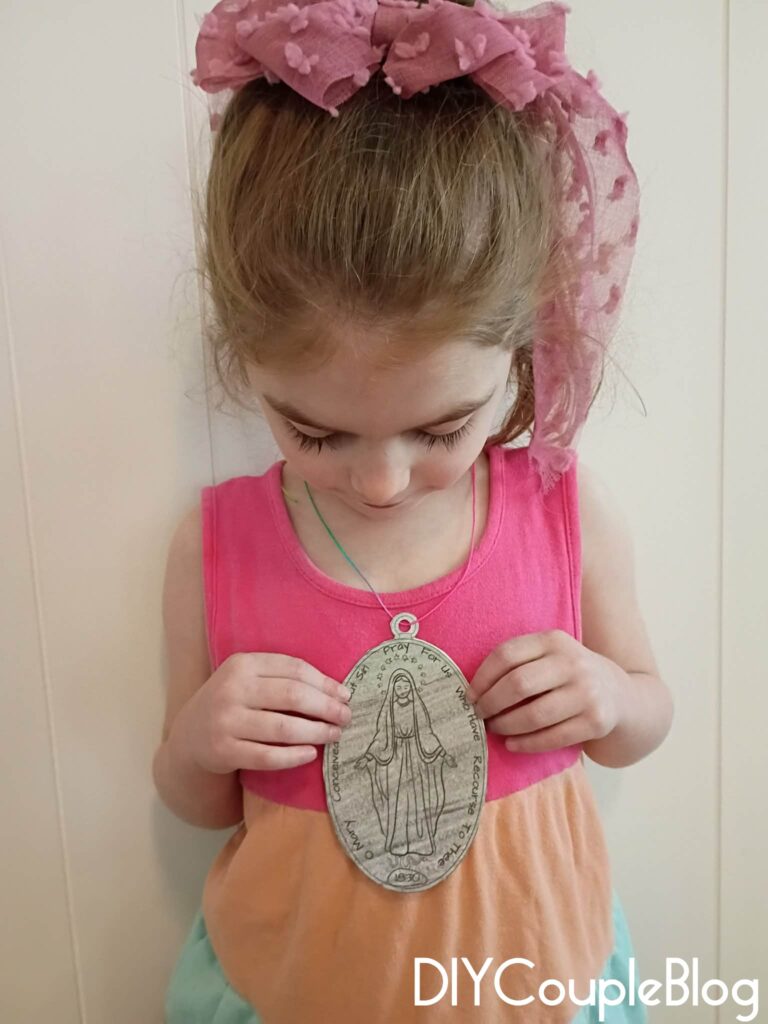 girl holding and showing the front a catholic printable miraculous medal craft necklace colored in "silver" crayon